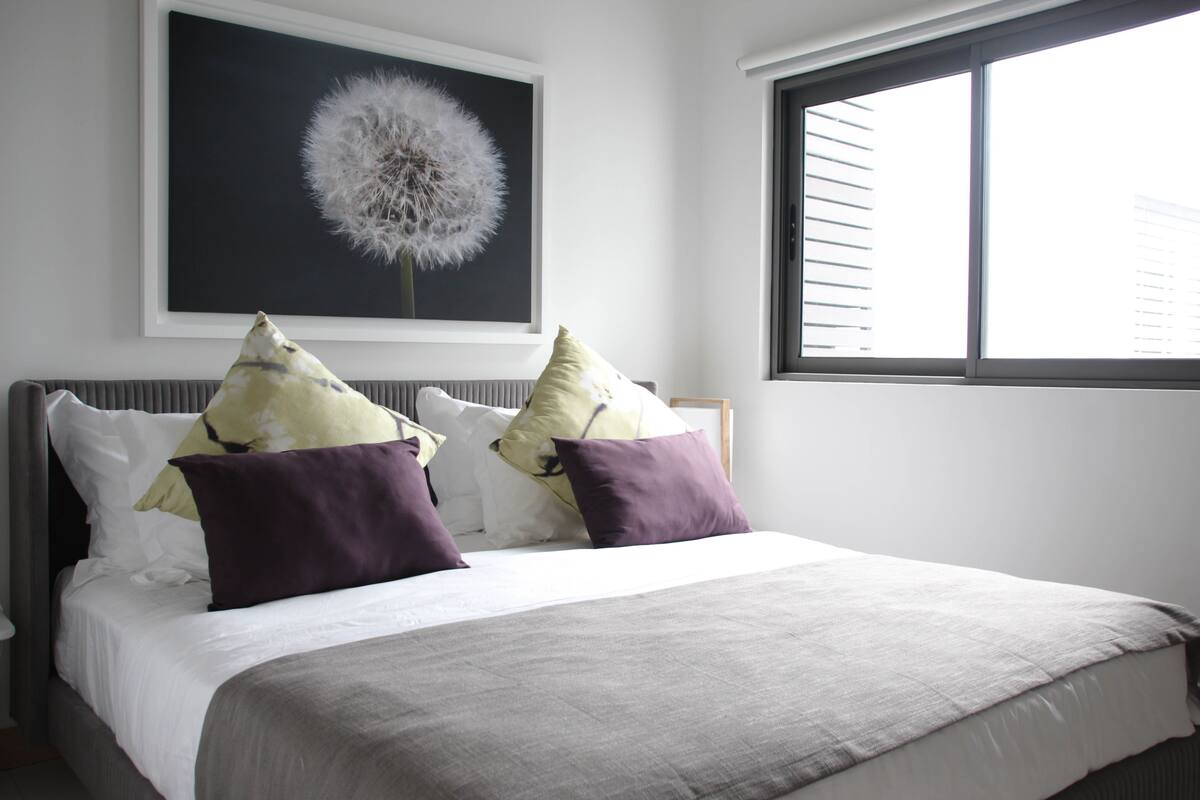 Una cama bien hecha con una colcha gris y varios almohadones, una obra de arte enmarcada de un diente de león en la pared, y una ventana con persianas.