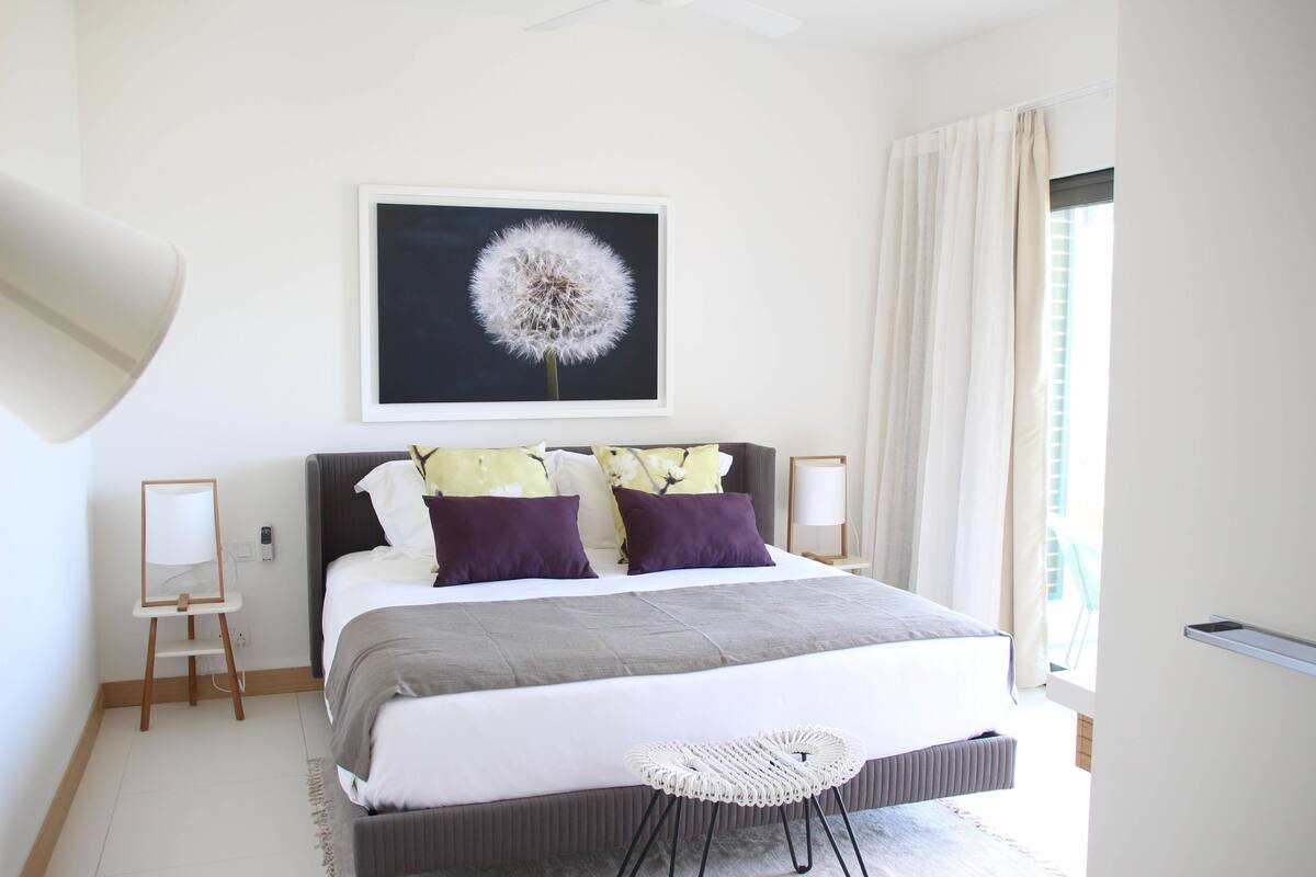 Un dormitorio moderno con una cama grande, una obra de arte enmarcada de un diente de león y vistas al exterior a través de una ventana.