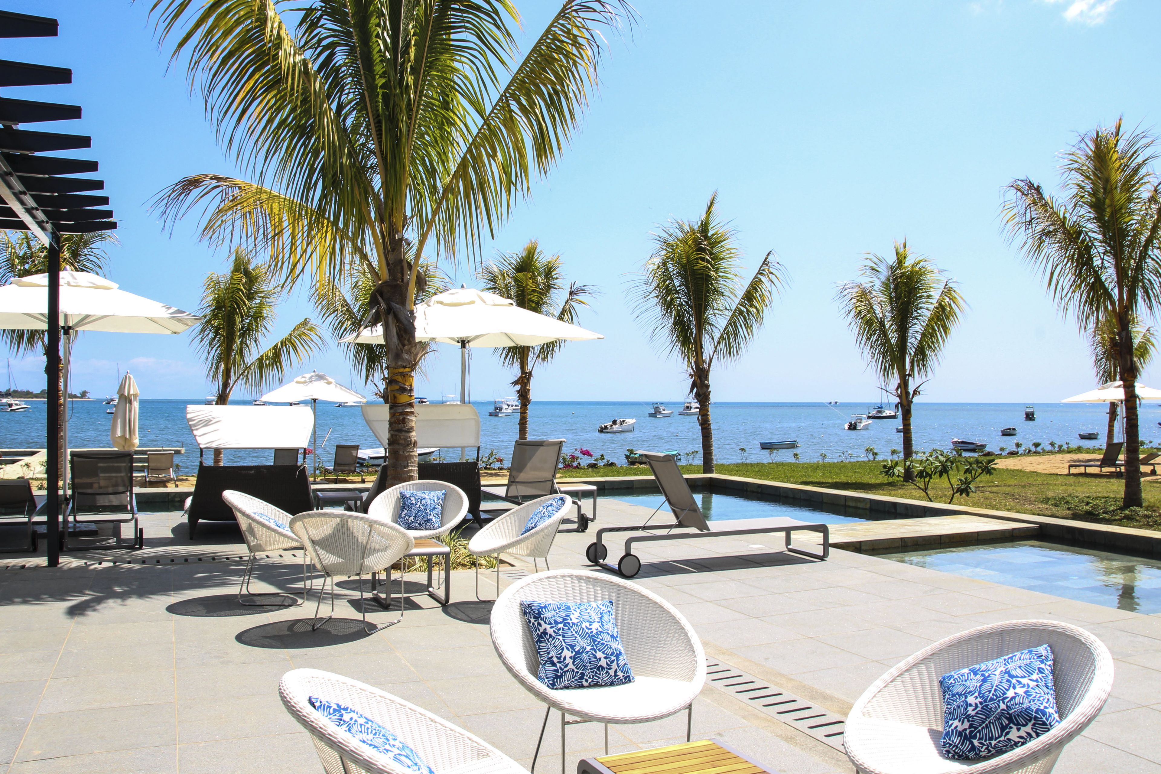 Un espace aménagé au bord de la piscine, avec des chaises longues et des parasols, donnant sur une plage où se trouvent des bateaux.