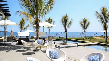 Un espace aménagé au bord de la piscine, avec des chaises longues et des parasols, donnant sur une plage où se trouvent des bateaux.