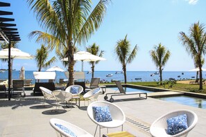 Área de piscina com espreguiçadeiras e guarda-sóis, com vista para uma praia com barcos.