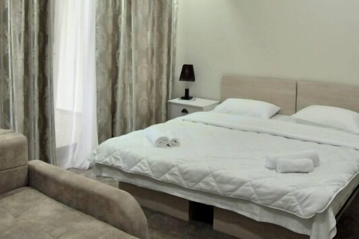 Una habitación de hotel con cama, mesita de noche, lámpara, sillón y cortinas.