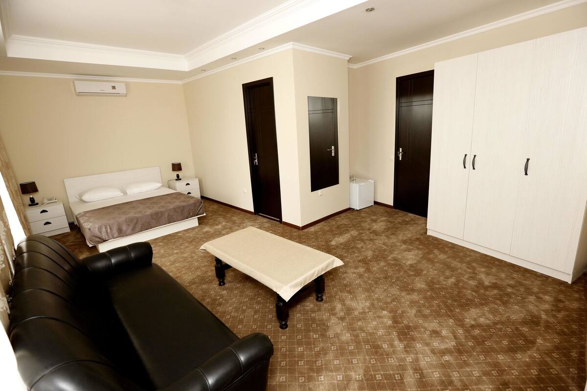 Habitación de hotel con cama, sofá de cuero negro, una mesita, armario y baño con inodoro y lavabo.