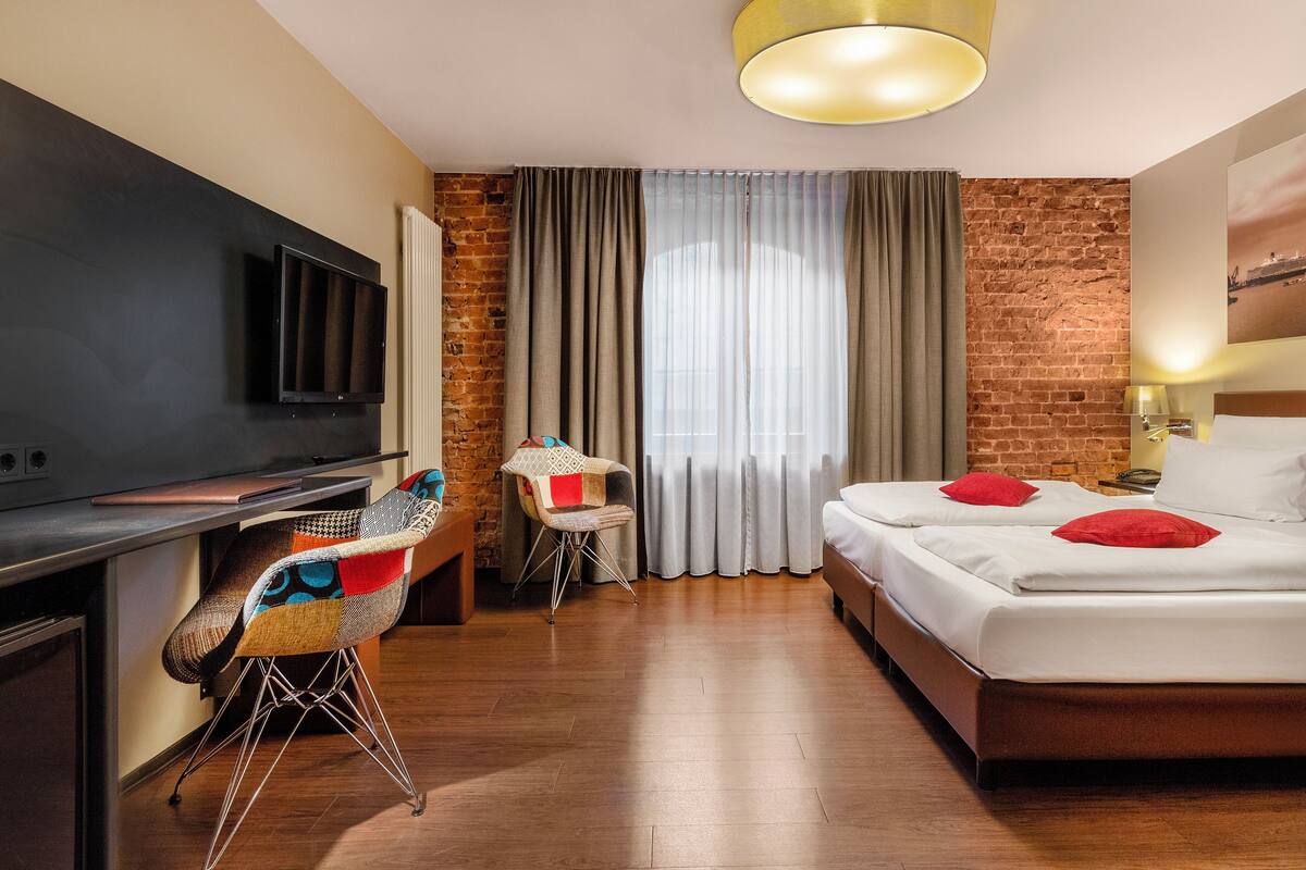 Habitación de hotel con cama, escritorio con televisor de pantalla plana, una silla y ventana con cortinas.