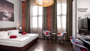 A hotel room with a bed, two chairs, a table, a TV, and a view of a park. - Hotel Volksschule (Hamburg)