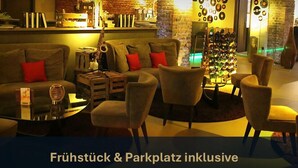A cozy lounge area with sofas and armchairs, a coffee table, and a wine rack. The text 'Frühstück & Parkplatz inklusive' is visible. - Hotel Volksschule (Hamburg)