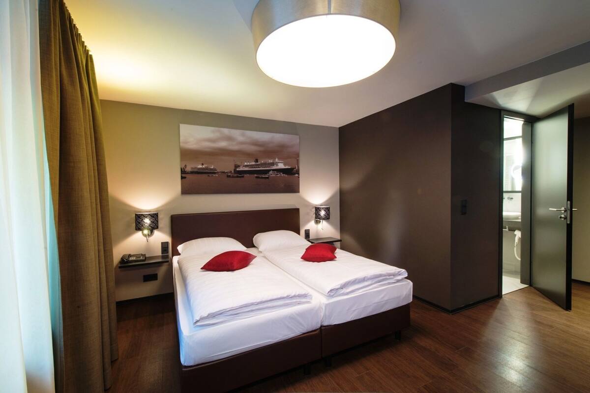 Una habitación de hotel con una cama doble, dos mesitas de noche, una lámpara de pared y un cuadro de un crucero.