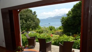 A balcony with wicker chairs, a table with fruit, and a view of the sea and mountains. - La Residence (Mahé Island)