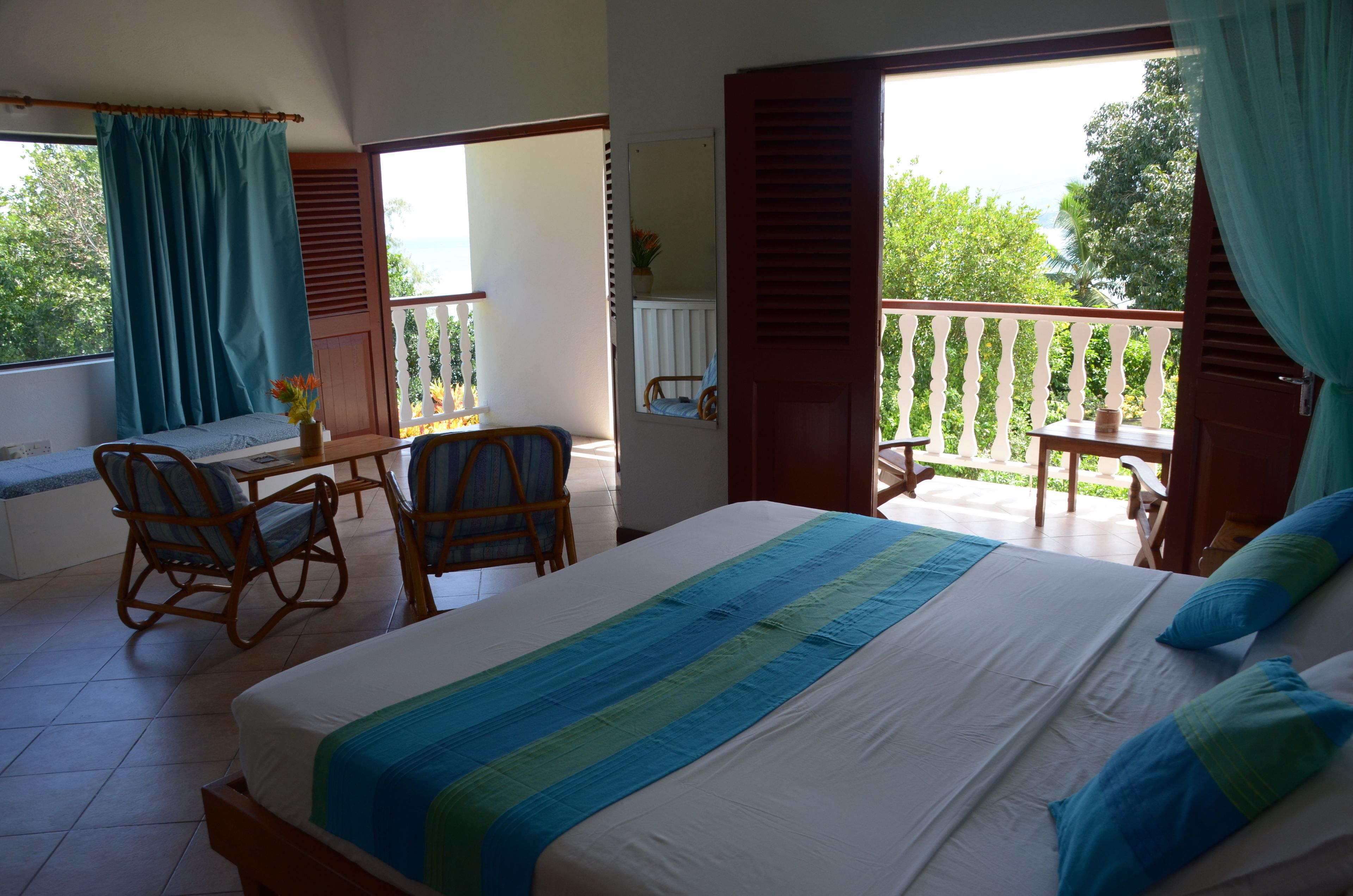 Une chambre à coucher avec un lit, un bureau et des chaises. Il y a un balcon d’où l’on peut admirer les arbres et le ciel.