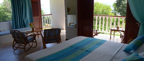 A bedroom with a bed, a desk, and chairs. There is a balcony with a view of trees and the sky.