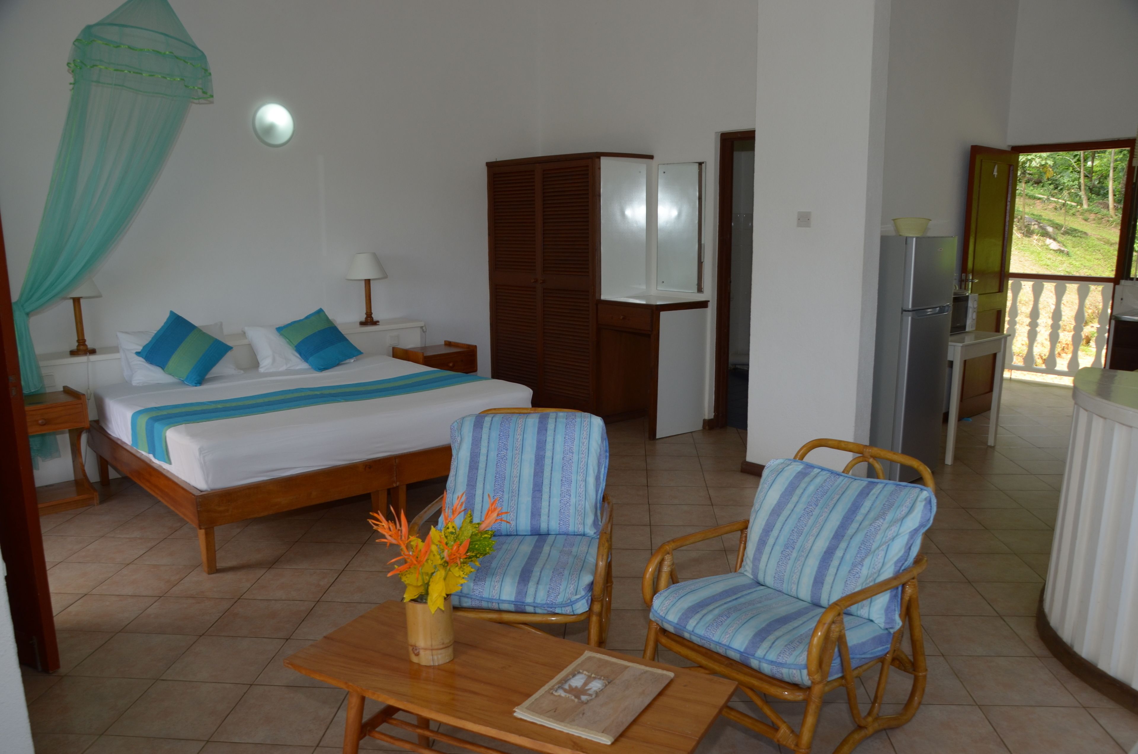 Un dormitorio con cama, mesitas de noche, un armario, una pequeña mesa con un florero, dos sillas de mimbre y vistas a un jardín a través de una puerta abierta.