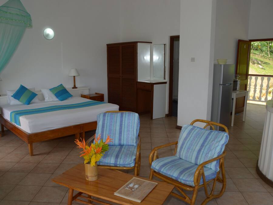 Un dormitorio con cama, mesitas de noche, un armario, una pequeña mesa con un florero, dos sillas de mimbre y vistas a un jardín a través de una puerta abierta.