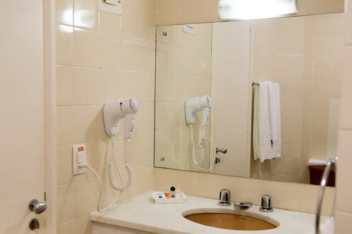 A bathroom with a sink, two hair dryers mounted on the wall, a mirror, and a towel hanging on the wall.