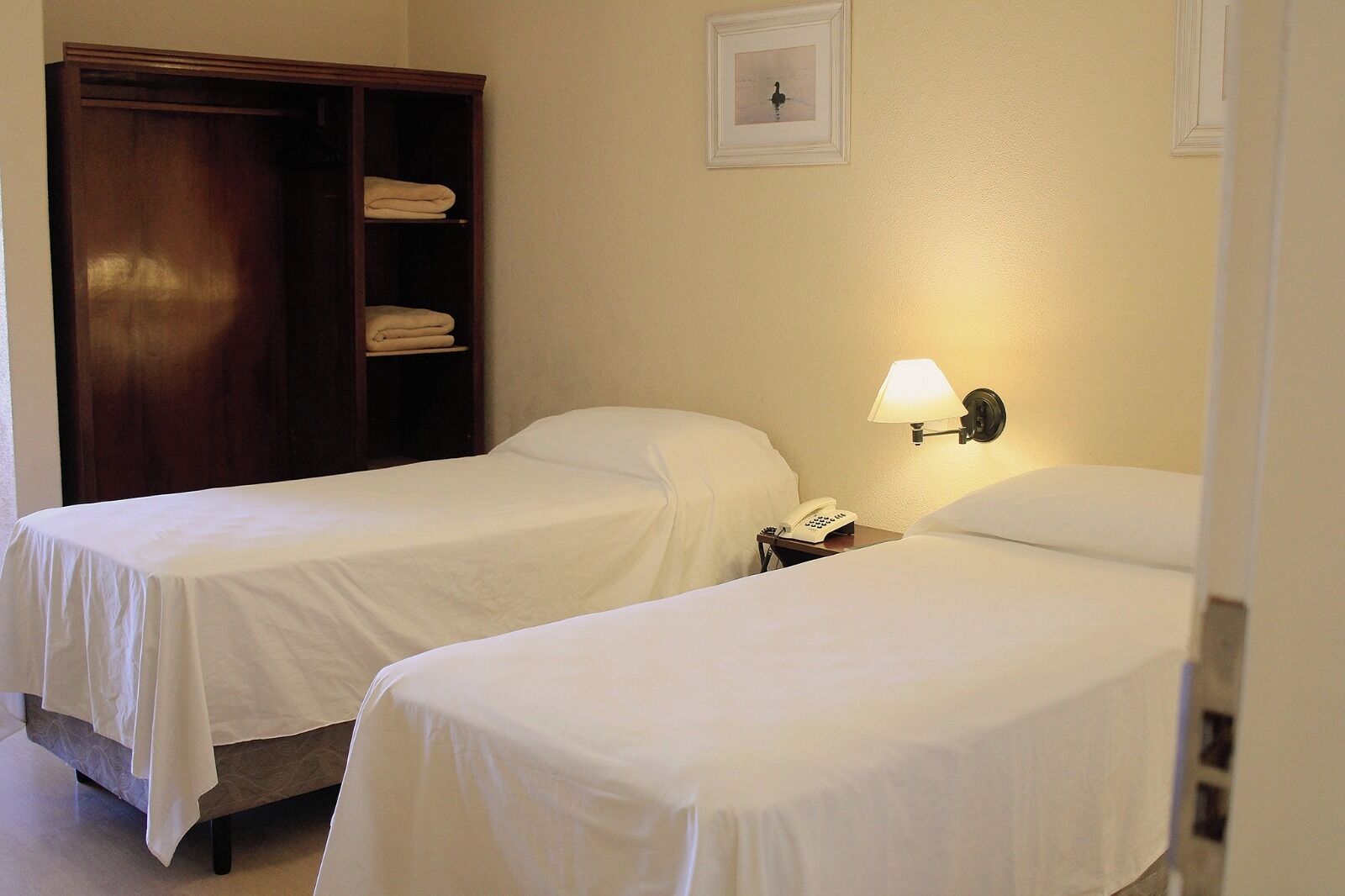 A hotel room with two single beds, a wardrobe, a bedside lamp, and a framed picture on the wall.