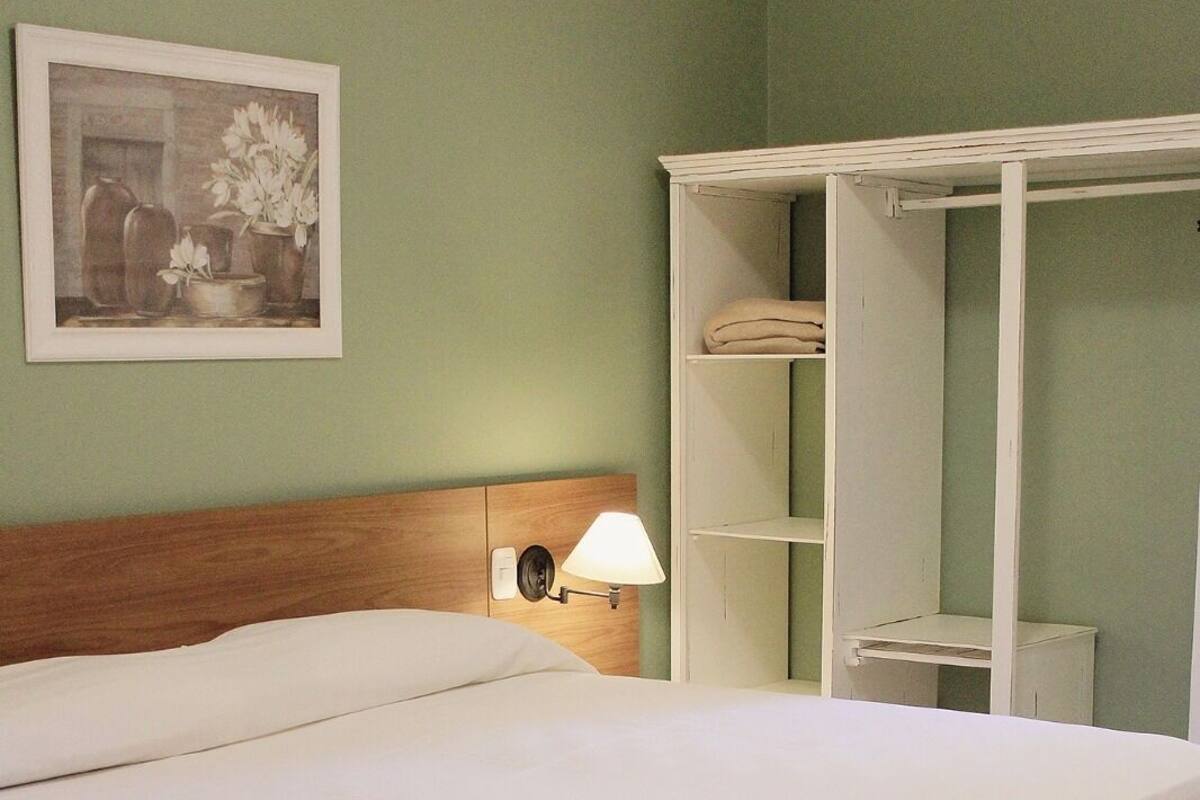 Un lit bien fait dans une pièce aux murs verts, avec un tableau encadré au-dessus du lit et une armoire blanche avec des étagères.