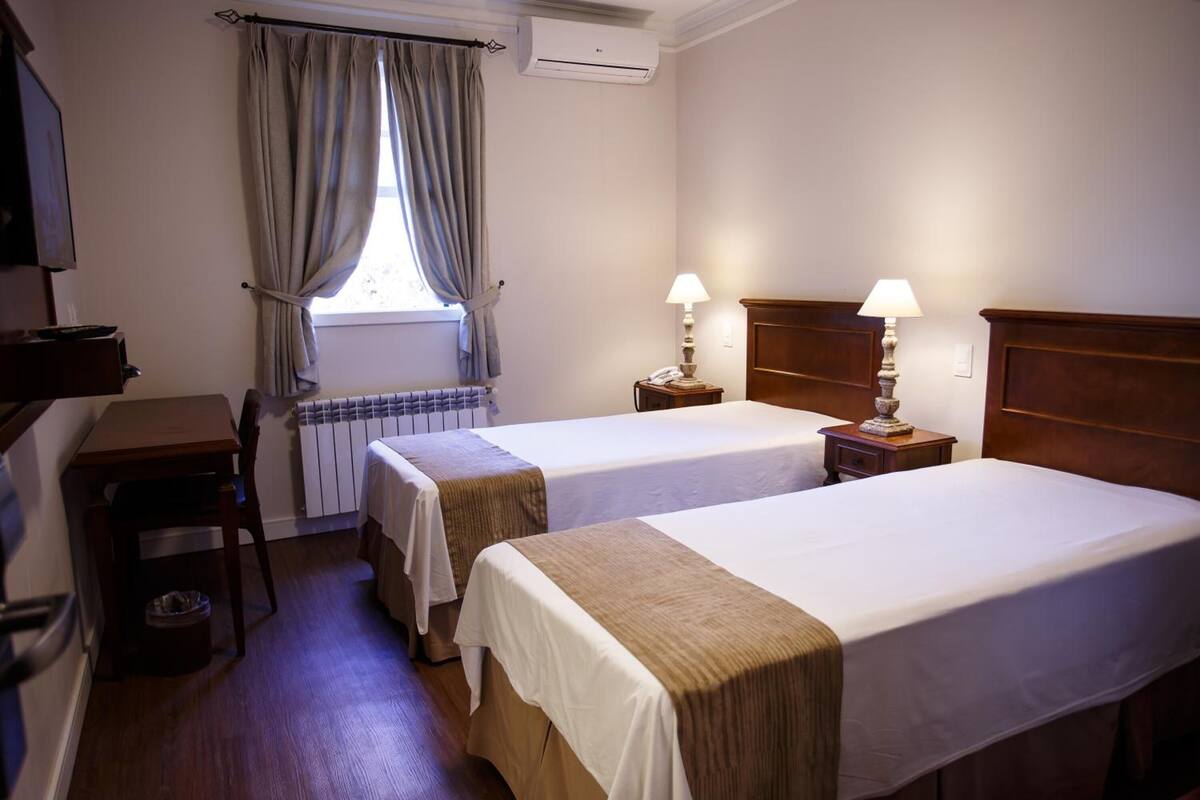 Une chambre d’hôtel avec deux lits, un bureau, une chaise, un radiateur, une fenêtre avec des rideaux et une lampe sur chaque table de chevet.