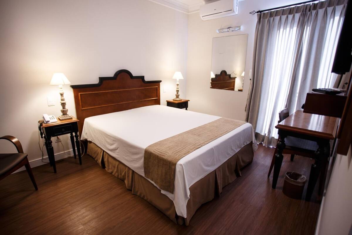 Une chambre d’hôtel avec un lit en bois, deux tables de chevet avec des lampes, une chaise, un bureau et un miroir au mur.
