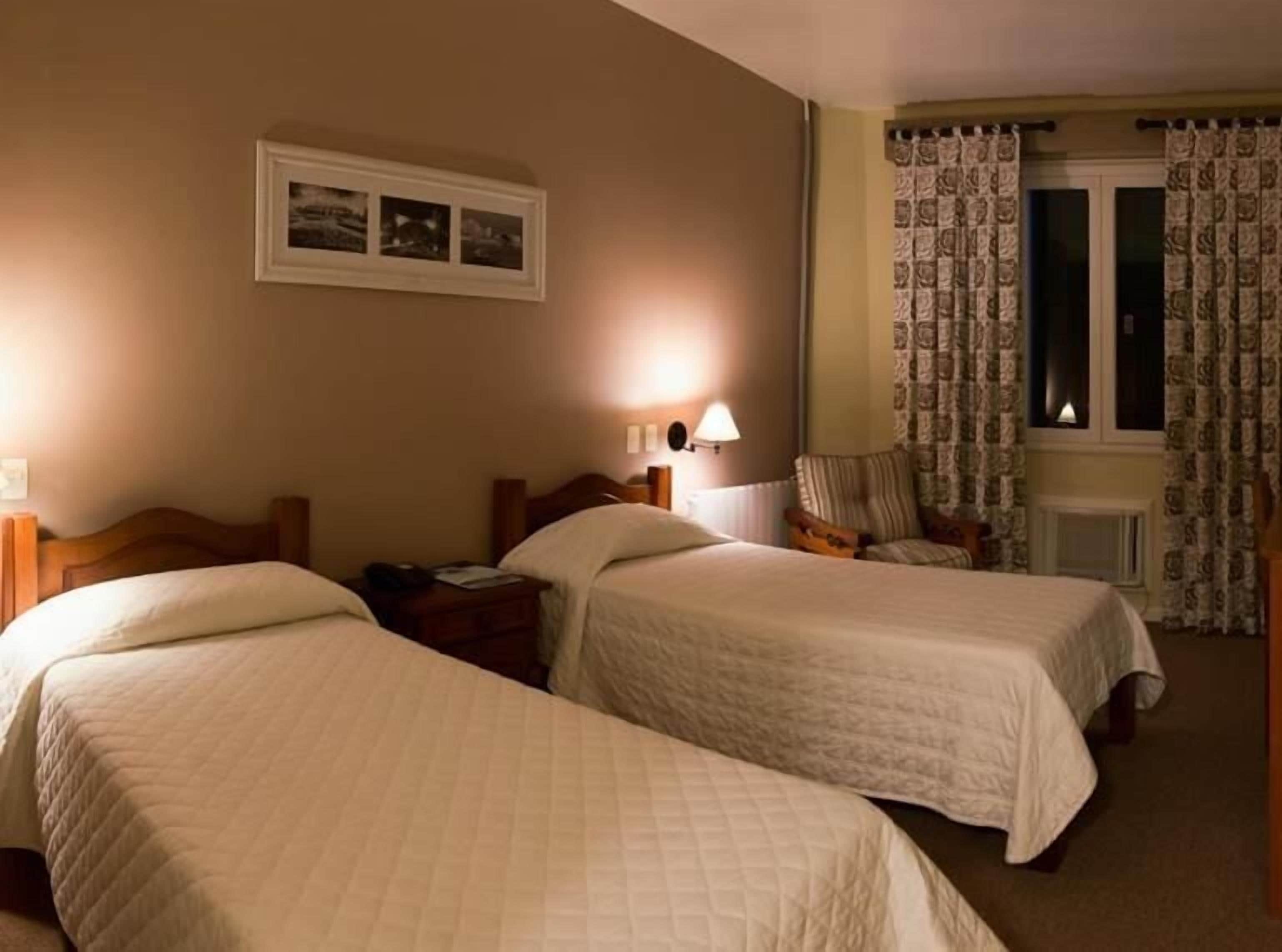 Habitación de hotel con dos camas individuales, una mesita de noche con lámpara, una silla y una ventana con cortinas estampadas.