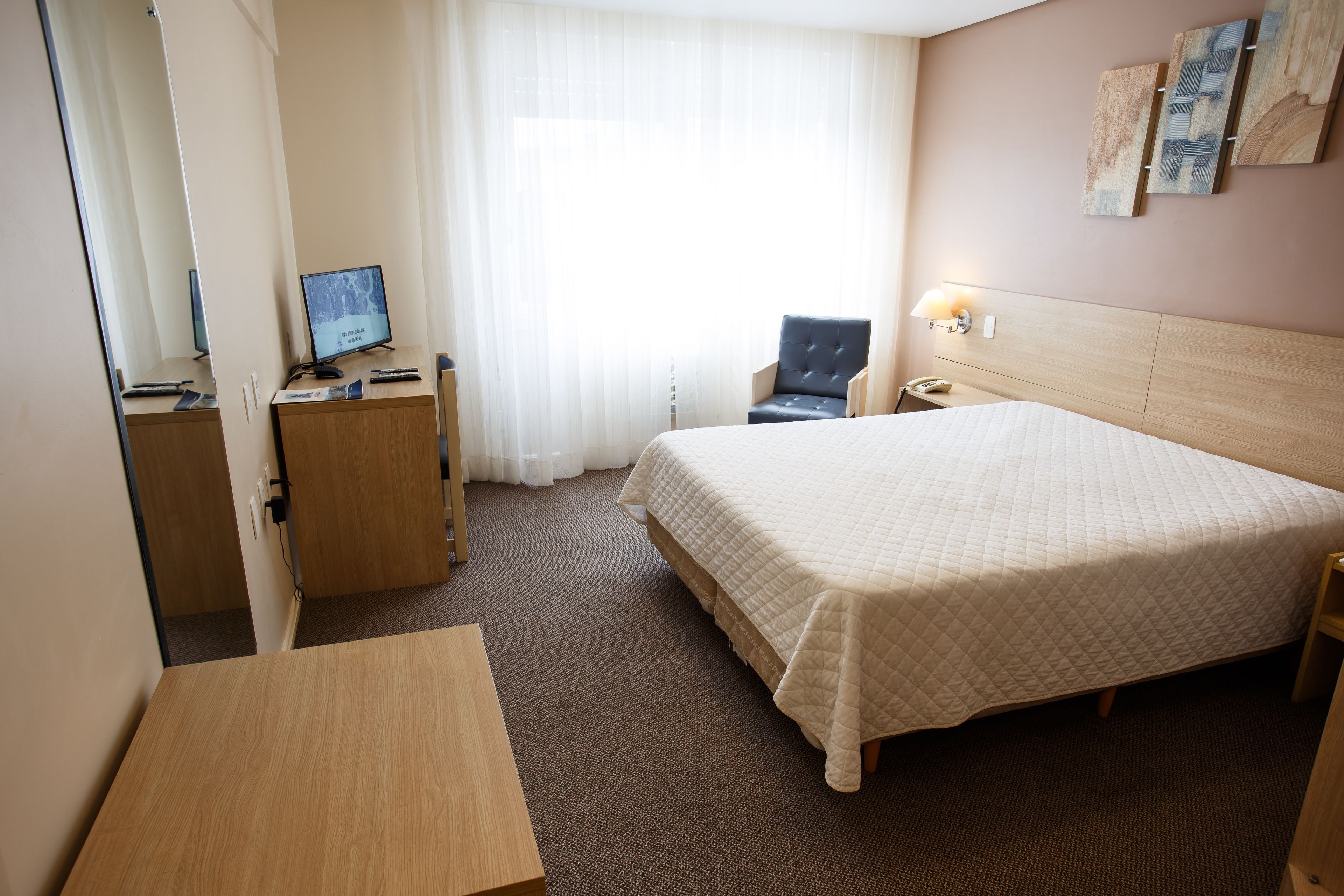 A hotel room with a bed, a desk, a chair, a TV, and a window with curtains.