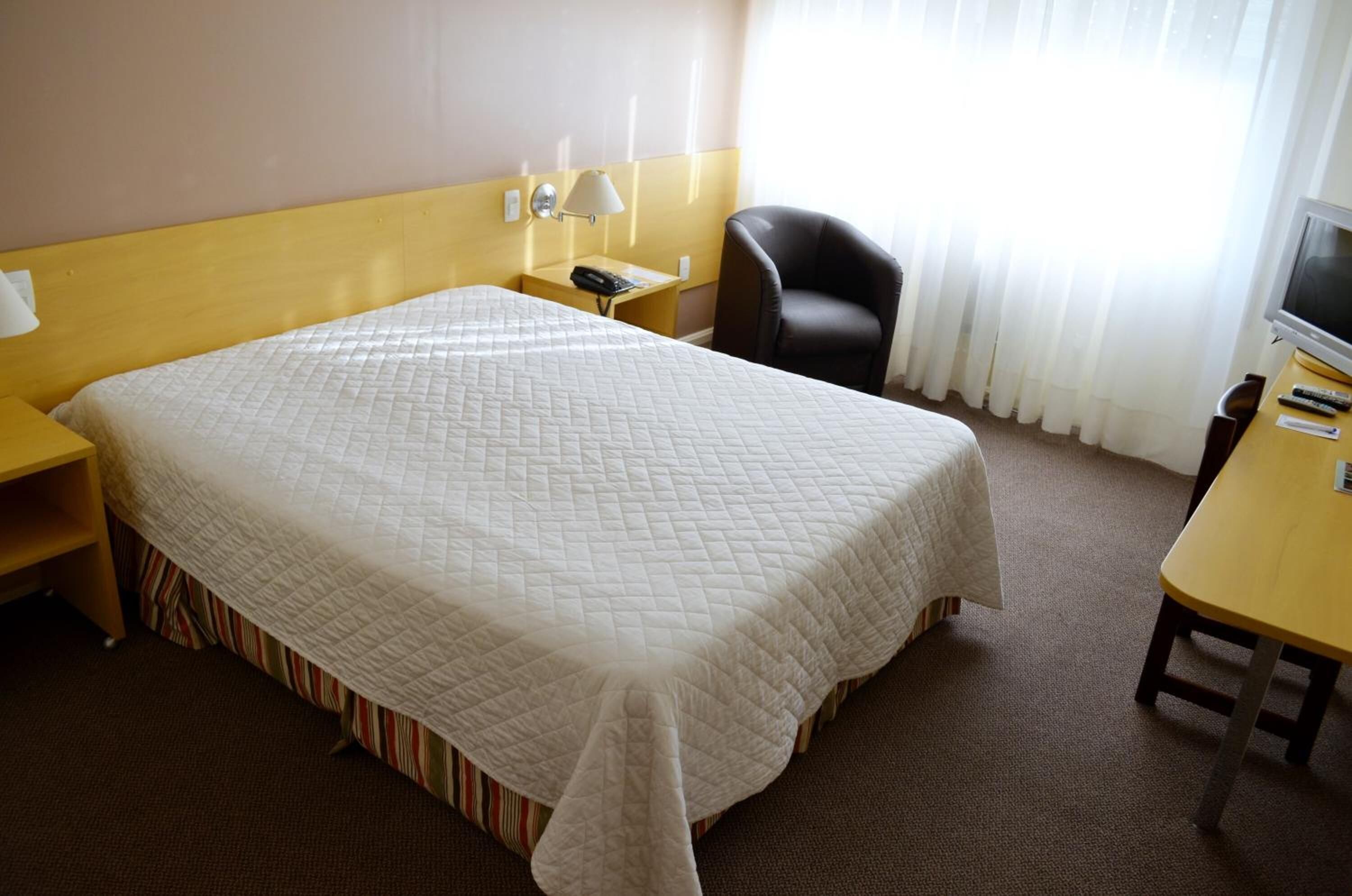 A hotel room with a large bed, a desk with a lamp, a chair, and a TV.