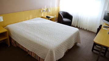 A hotel room with a large bed, a desk with a lamp, a chair, and a TV.