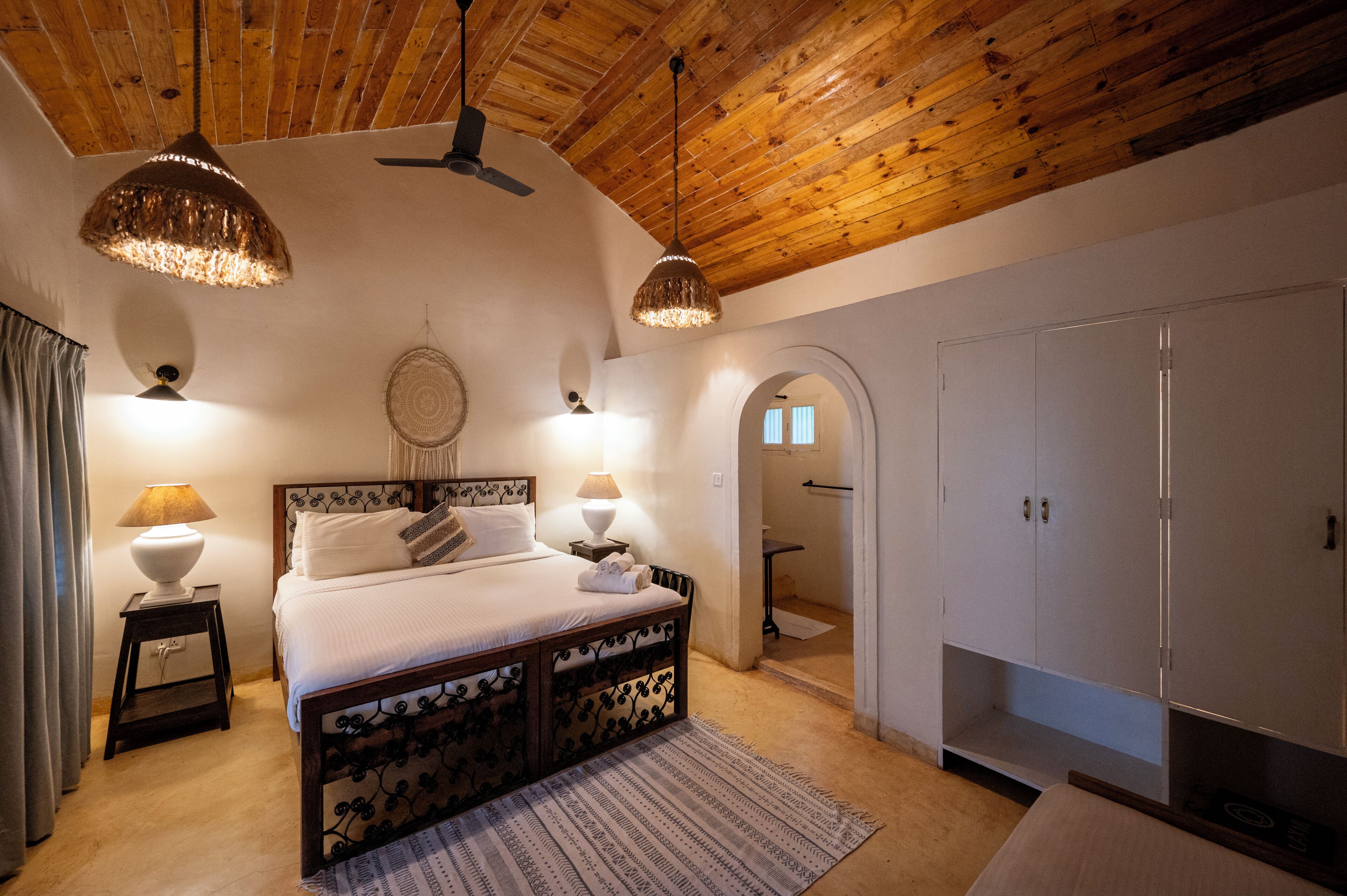 A bedroom with a wooden ceiling, a bed with a headboard, two bedside lamps, a wall-mounted dreamcatcher, and a wardrobe.