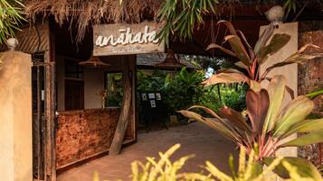 Der Eingang zu einem rustikalen Rückzugsort mit einem Strohdach, einer Holztür und einem Schild mit der Aufschrift 'Anikato Seaside Retreat'.