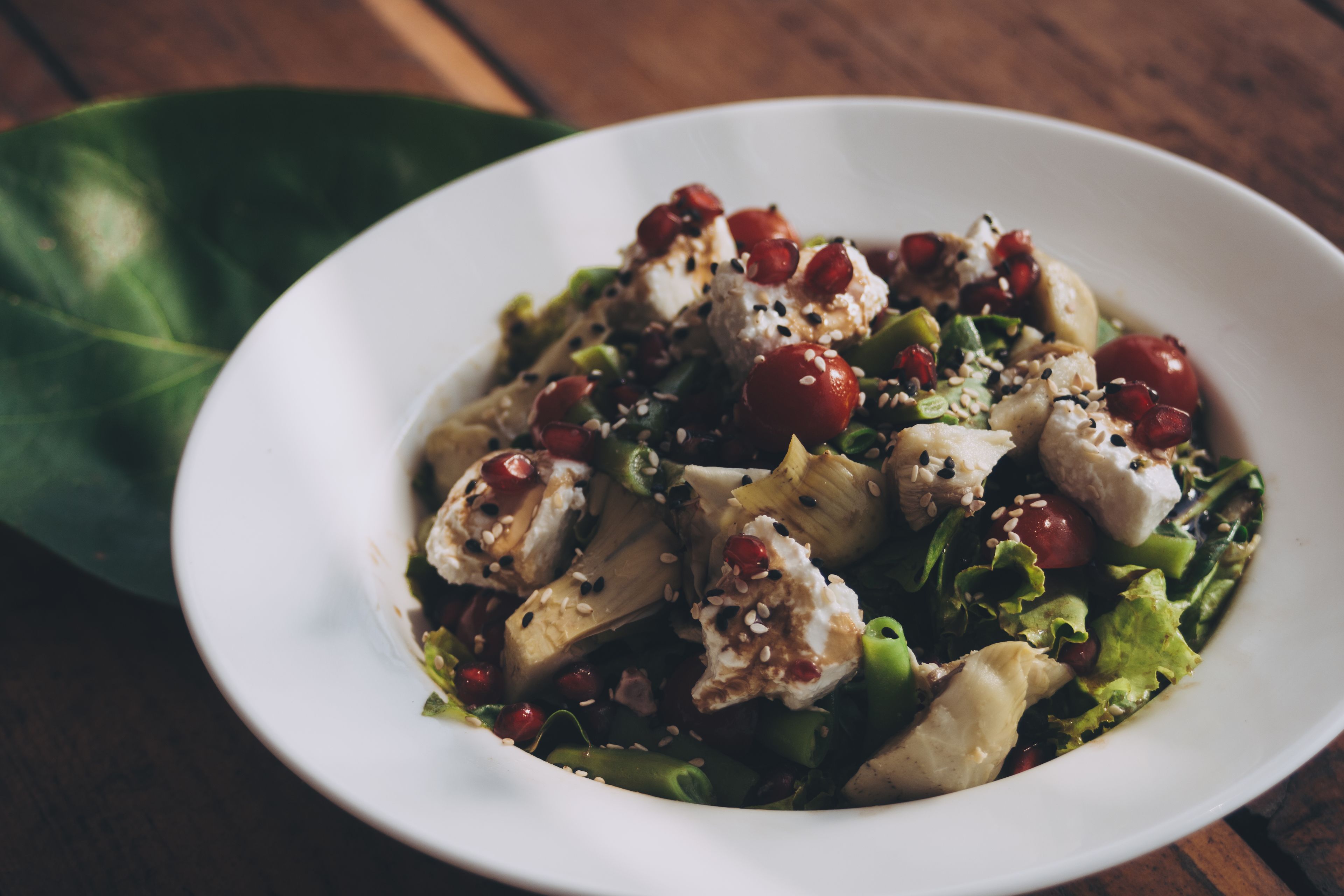 A white bowl of salad with various ingredients, including greens, tomatoes, and sesame seeds, on a wooden table.