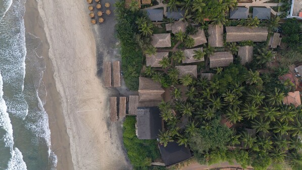Vista aérea de una propiedad frente a la playa con construcciones de techo de paja, palmeras y una playa arenosa.