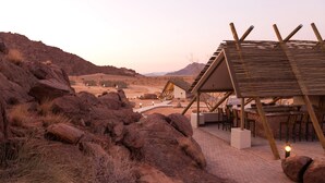 A rustic outdoor seating area with wooden tables and chairs, surrounded by large rocks and a desert landscape in the background. - Desert Quiver Camp (Sesriem)