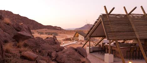 A rustic outdoor seating area with wooden tables and chairs, surrounded by large rocks and a desert landscape in the background.