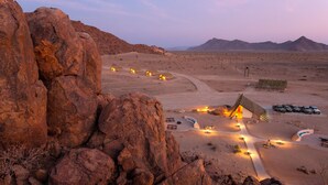 A desert landscape with a large rock formation in the foreground, a building with a sloped roof, and several vehicles parked nearby. - Desert Quiver Camp (Sesriem)