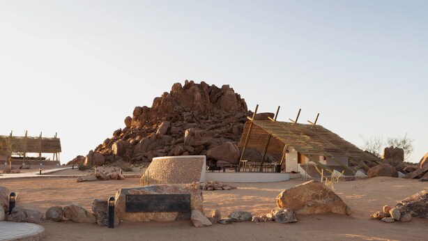 A desert camp with a large rock formation, a thatched-roof structure, and a sign reading "Desert Oasis Camp". - Desert Quiver Camp (Sesriem)