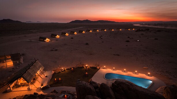 A desert resort with multiple lodges, a swimming pool, and a large rock formation in the foreground. - Desert Quiver Camp (Sesriem)