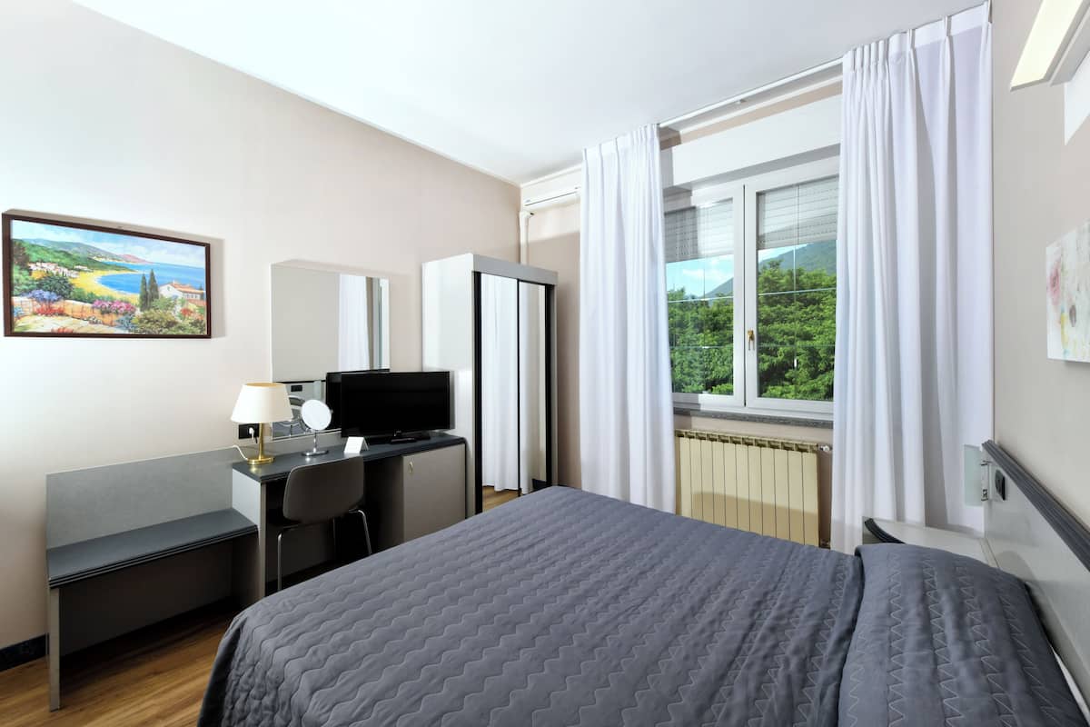 A hotel room with a bed, a desk with a computer, a chair, a TV, and a window with curtains.