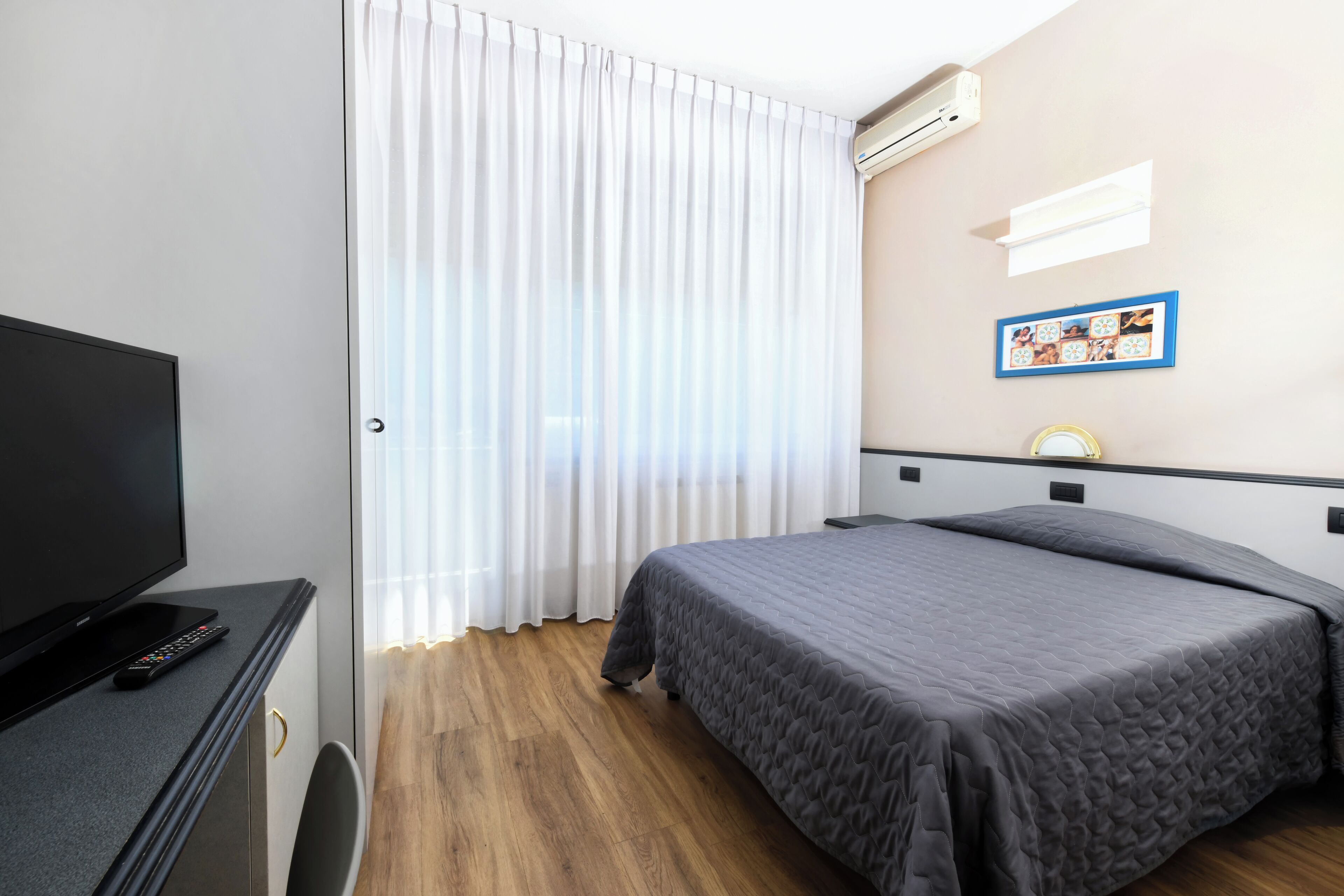 Una camera d'albergo con un letto, una televisione, un telecomando, una finestra con tende, un condizionatore d'aria e un quadro appeso al muro.