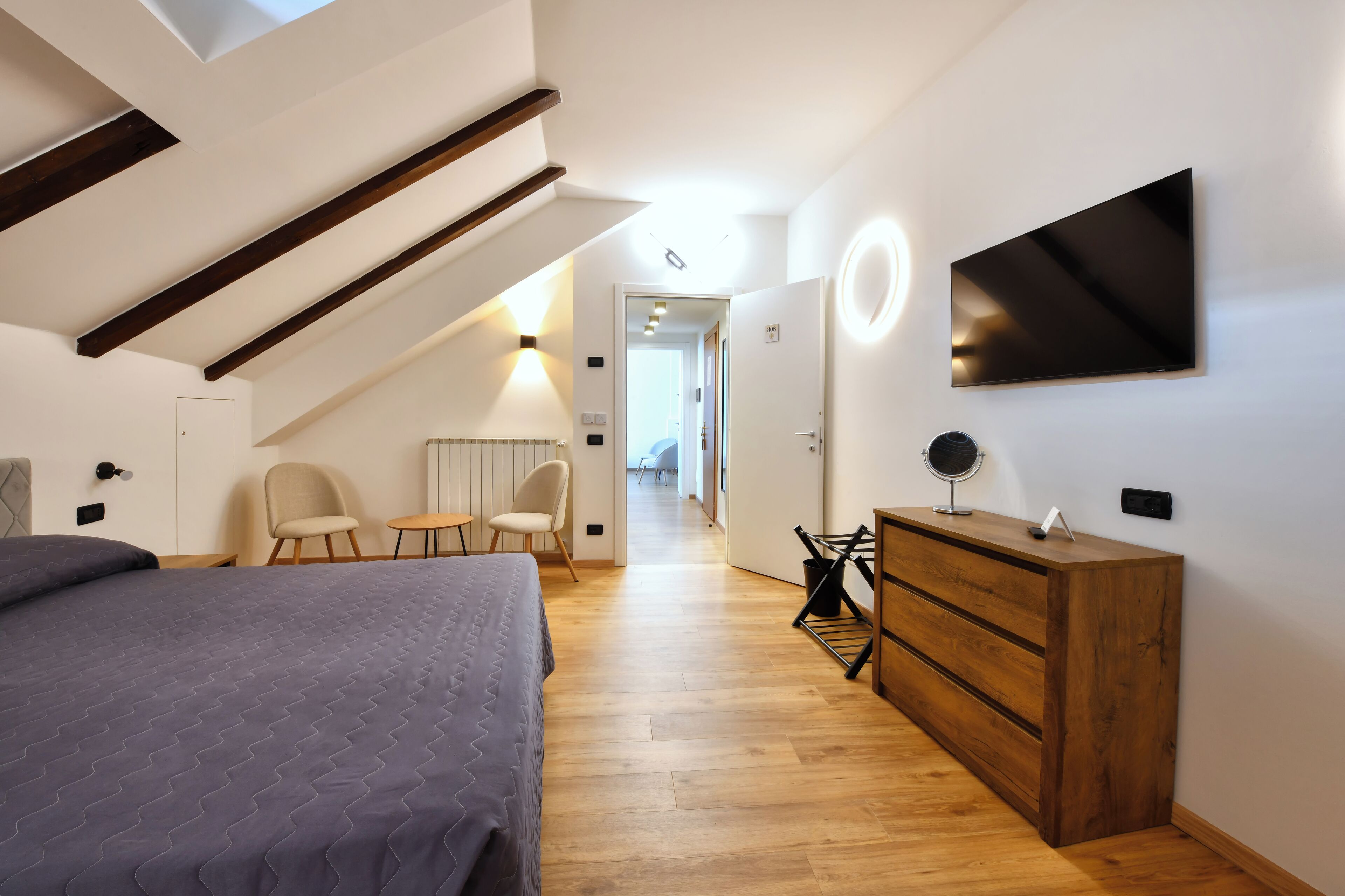 A modern bedroom with a sloped ceiling, a bed, a wooden dresser, a TV, and a desk with chairs.