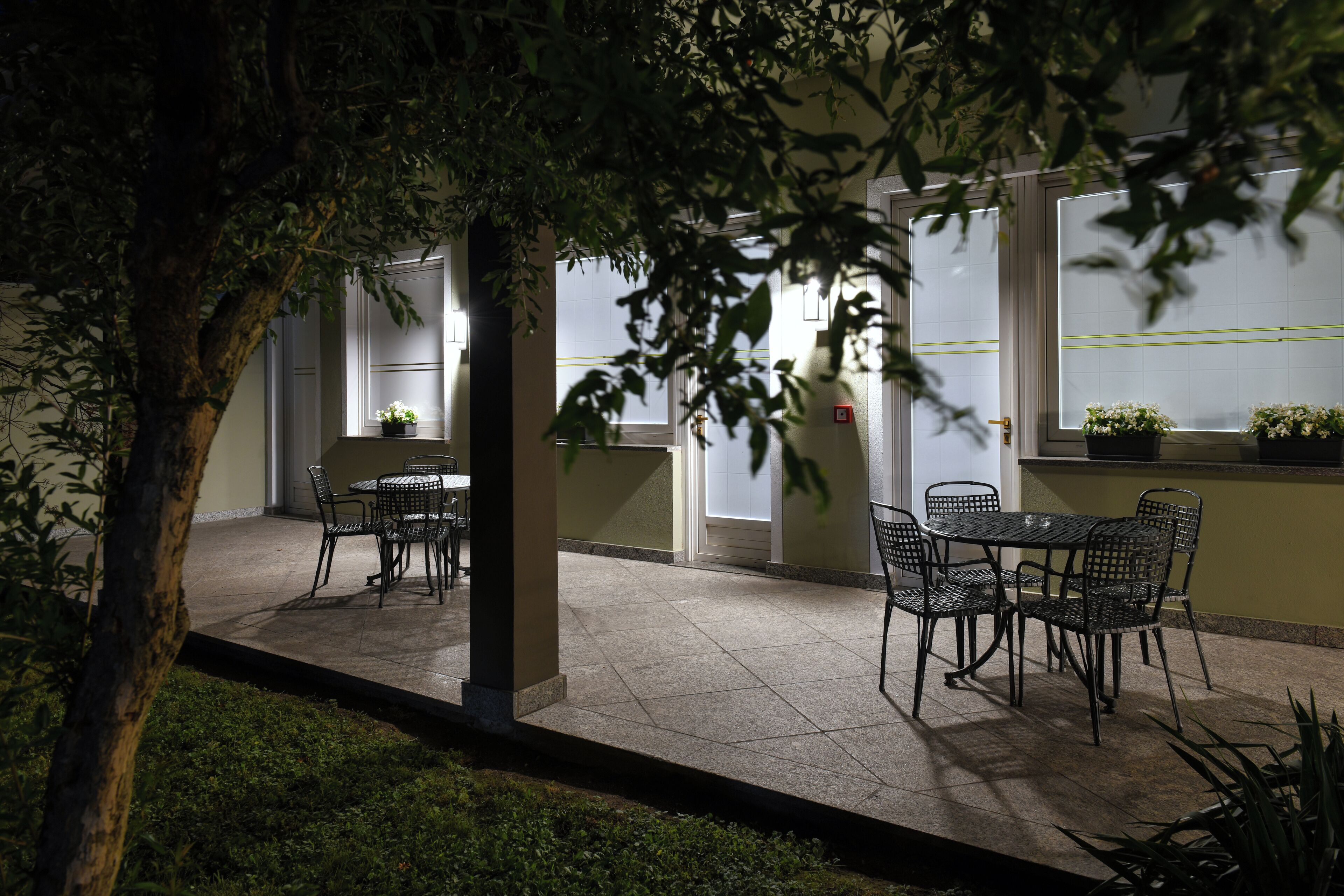 An outdoor patio area with tables and chairs, surrounded by greenery and a building with large windows.