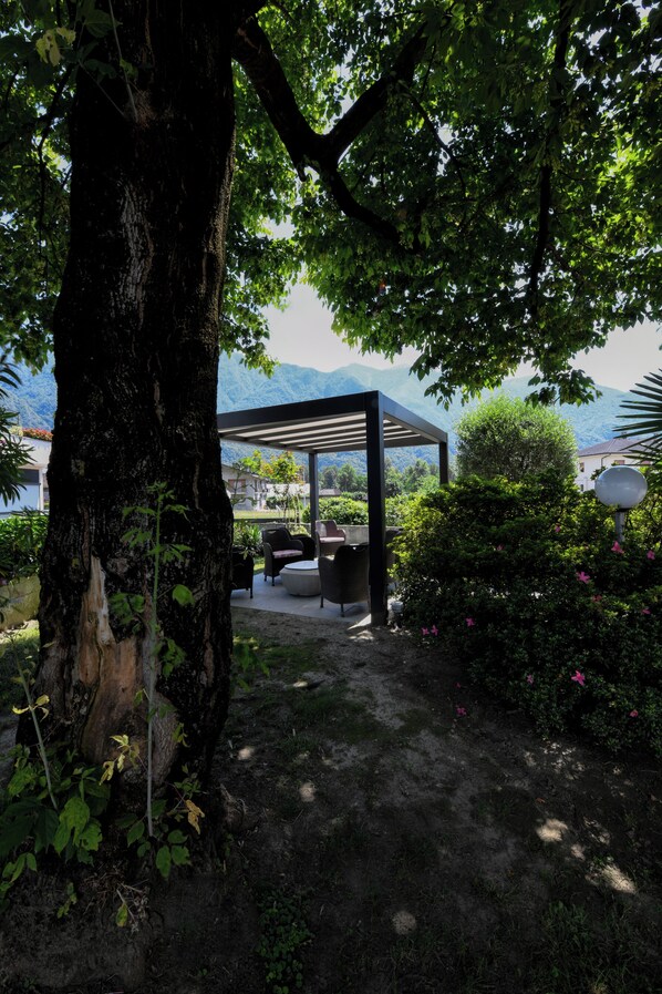 A covered outdoor seating area with a pergola, surrounded by lush greenery and a view of mountains. - Hotel Internazionale (Domodossola)