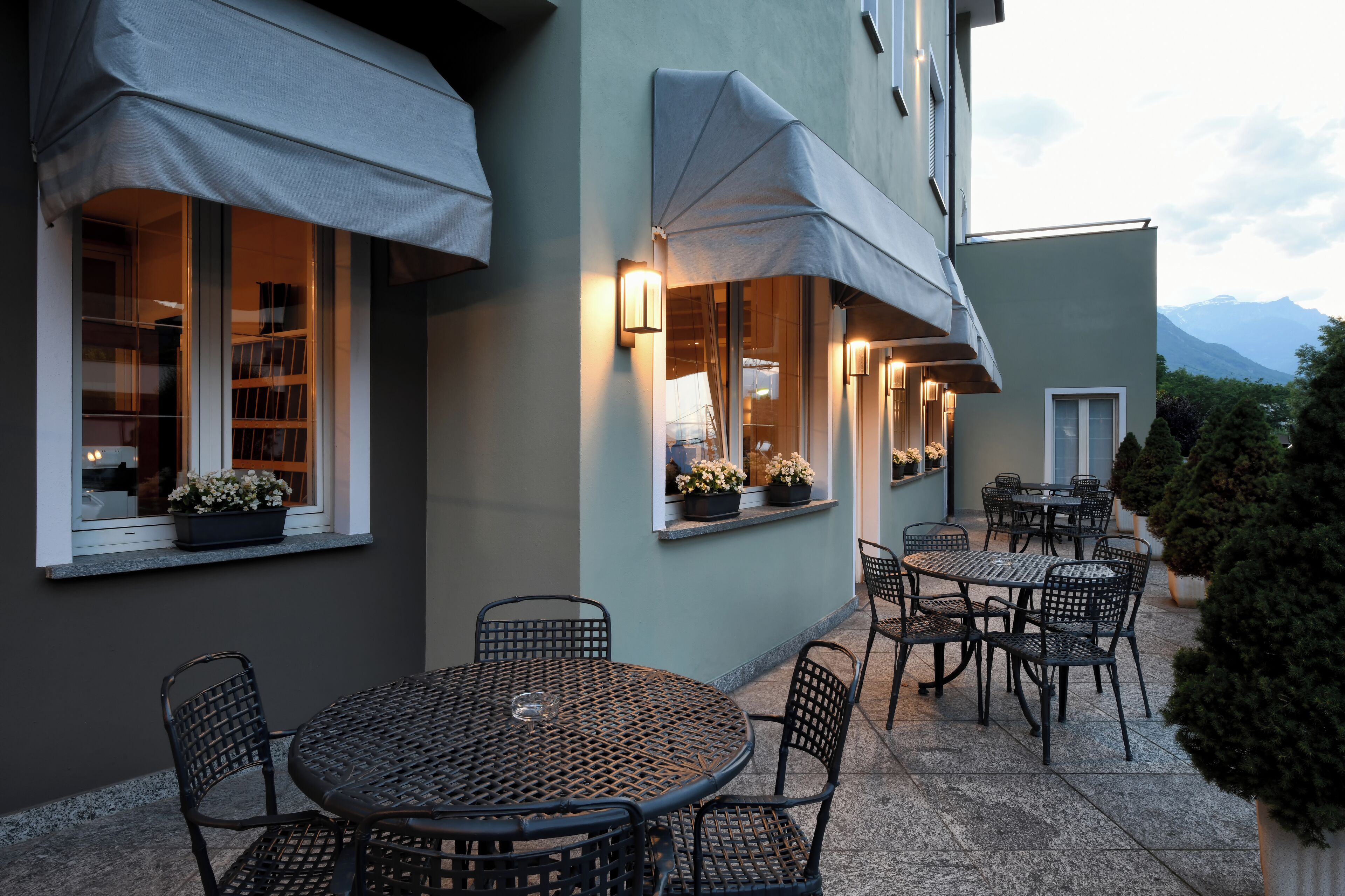 Ein Terrassenbereich im Freien mit runden Tischen und Stühlen, beschattet durch Sonnenschirme, mit Blumentöpfen an den Fensterbänken.