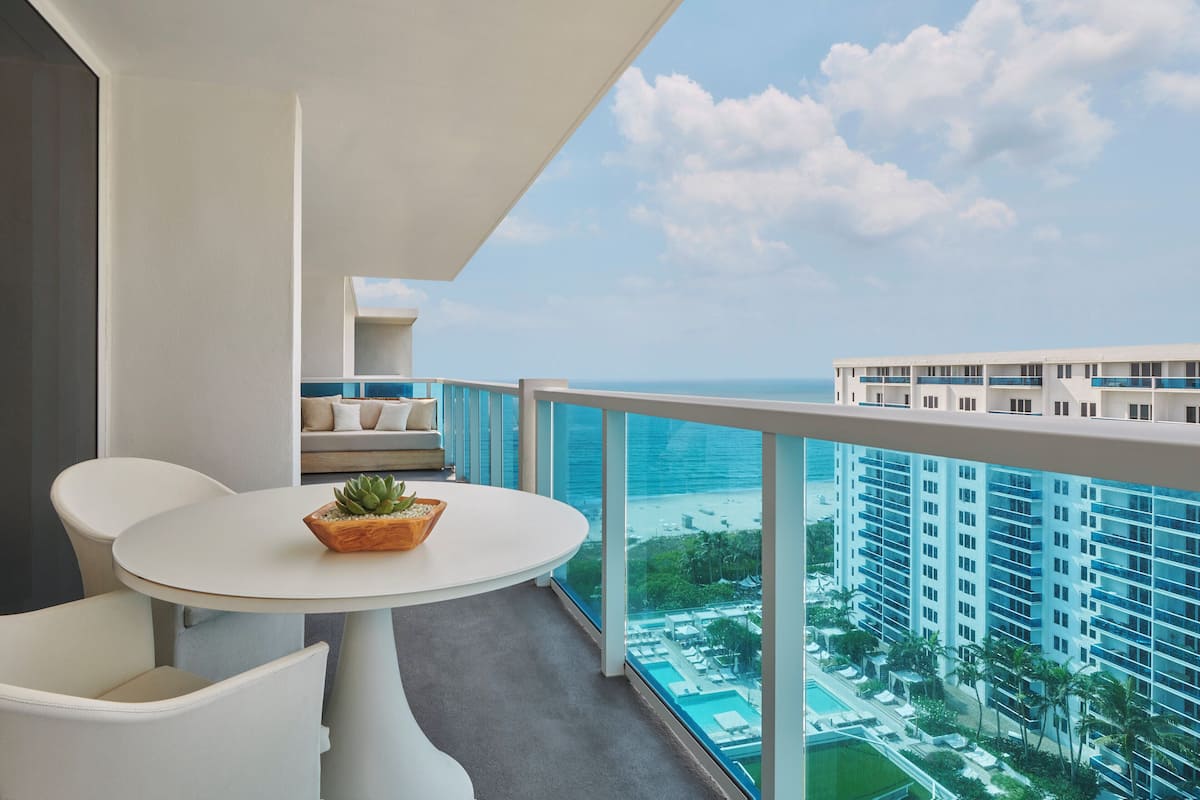 A balcony with a round table and chairs, a potted plant, and a view of a hotel building and beach.
