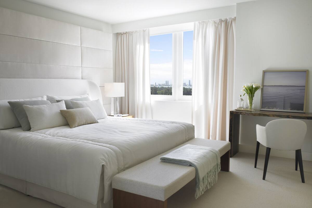 A modern hotel room with a large bed, a desk, a chair, and a view of the city through the window.