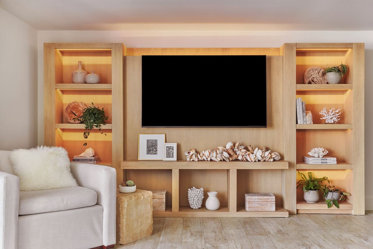 A modern living room with a flat-screen TV mounted on a wooden wall, surrounded by shelves with decorative items and plants.