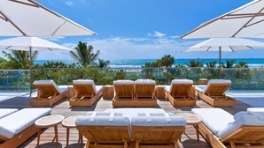 A rooftop lounge area with wooden loungers and white cushions, overlooking a beach with palm trees and the ocean.