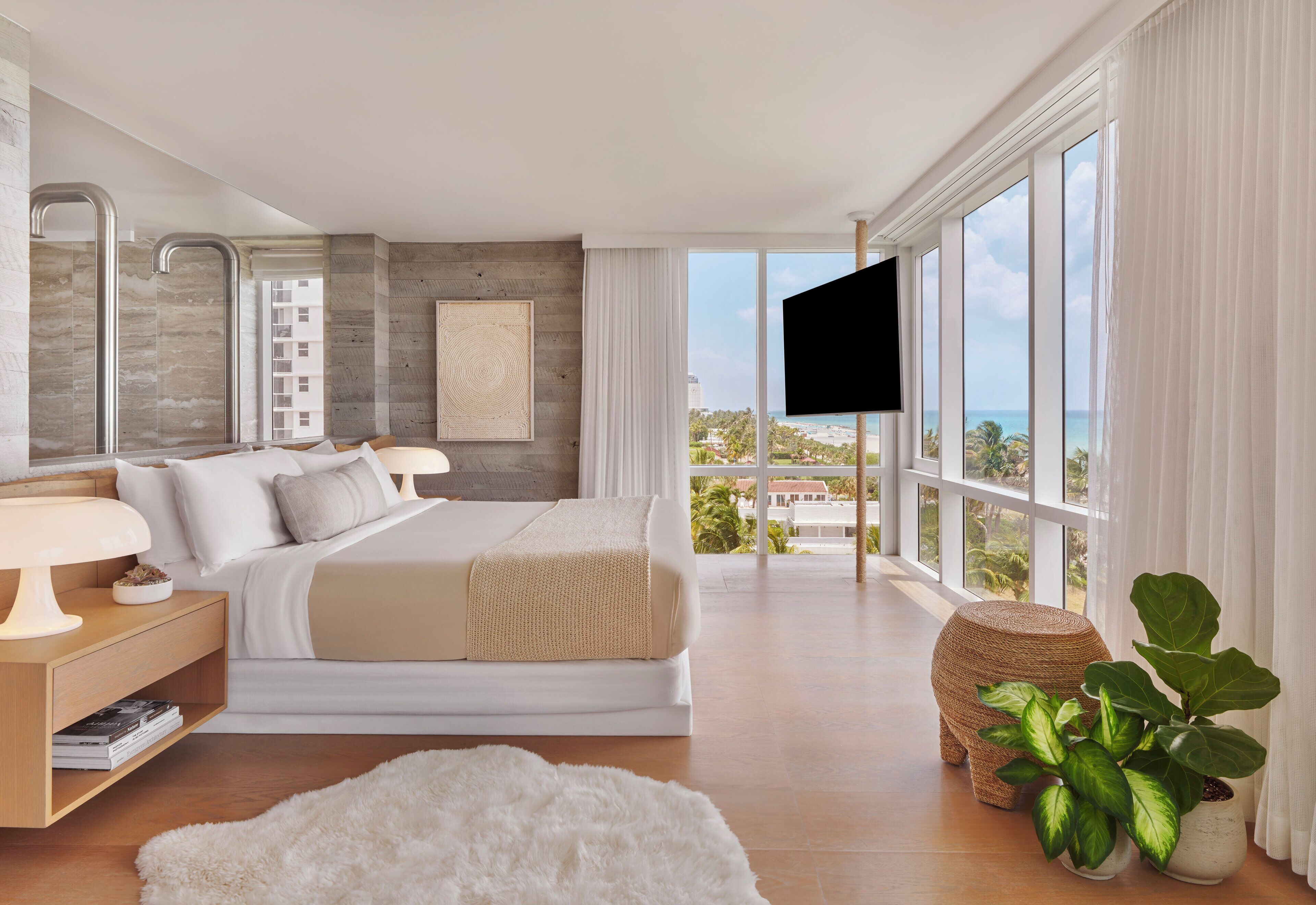 A modern bedroom with a large bed, a flat-screen TV, a view of the ocean, and a wooden floor.