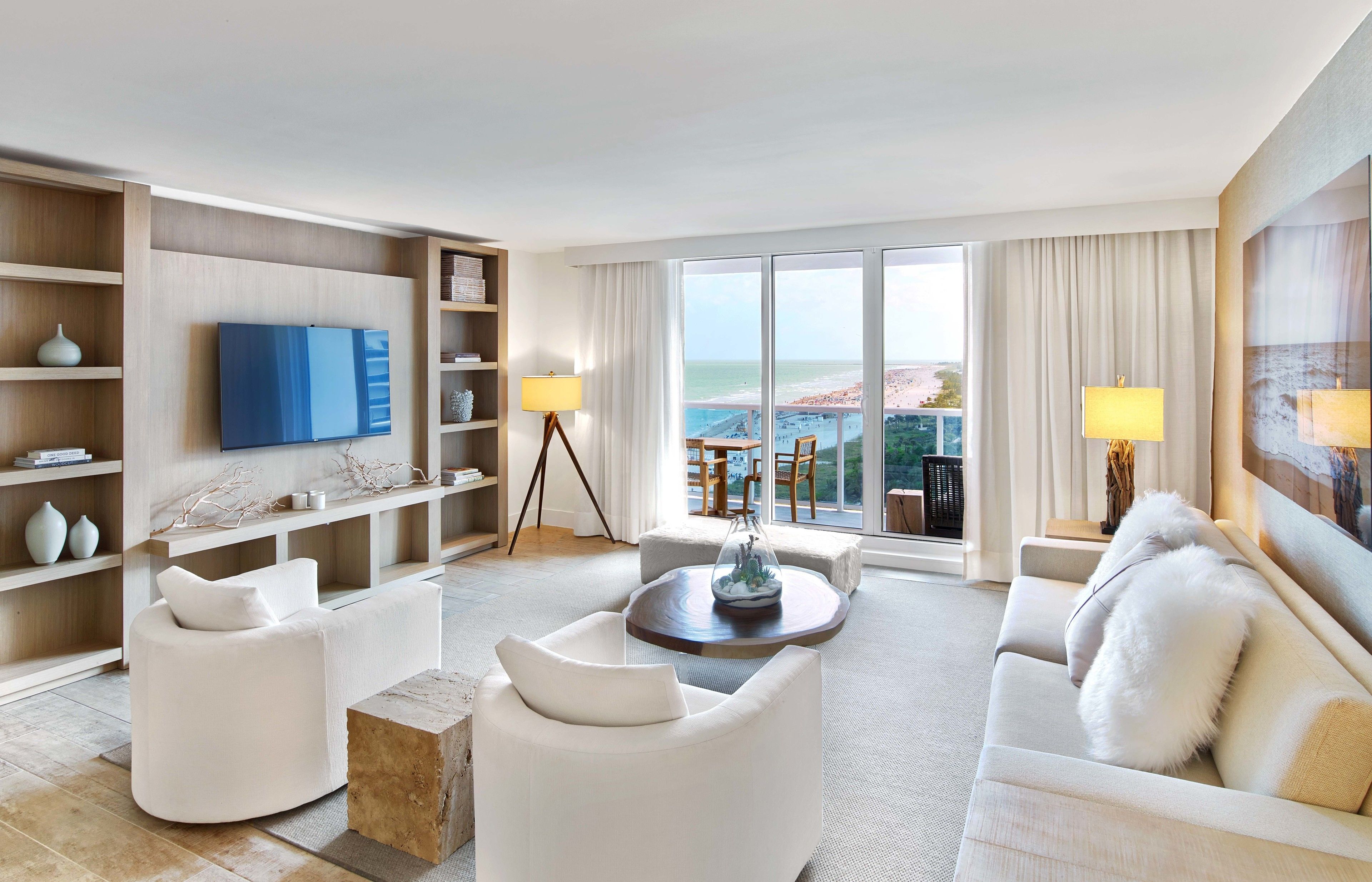 A modern hotel room with a large flat-screen TV, comfortable seating, a dining area, and a balcony with a view of the beach.