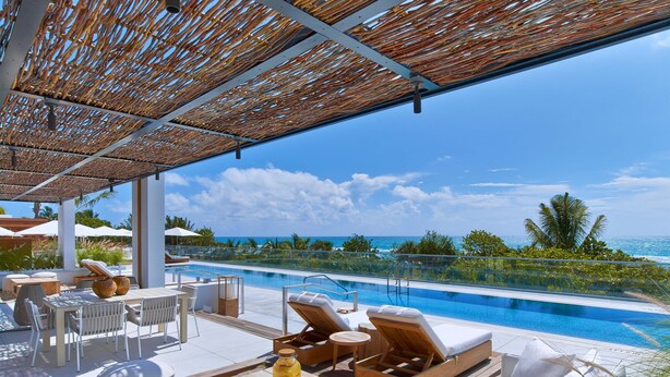Zona junto a la piscina con sillones de descanso, una mesa con bebida y vista al océano.