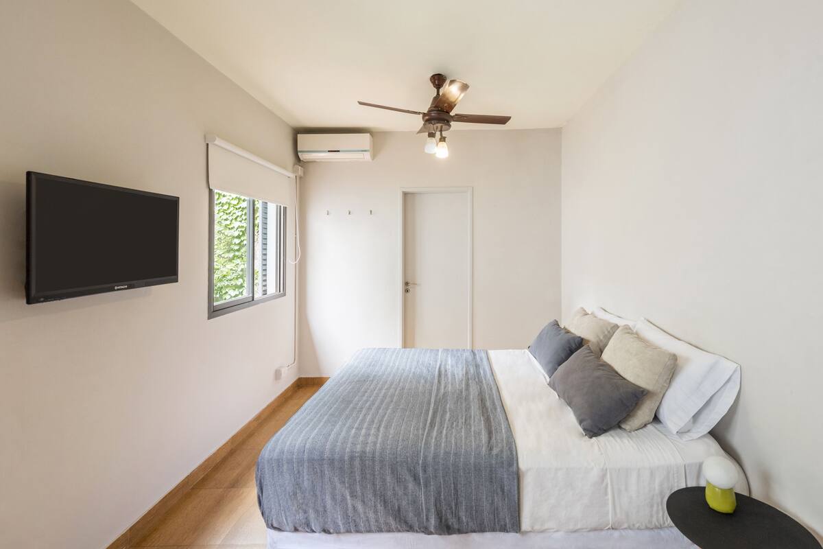 A bedroom with a bed, a ceiling fan, a TV mounted on the wall, and a window with blinds.