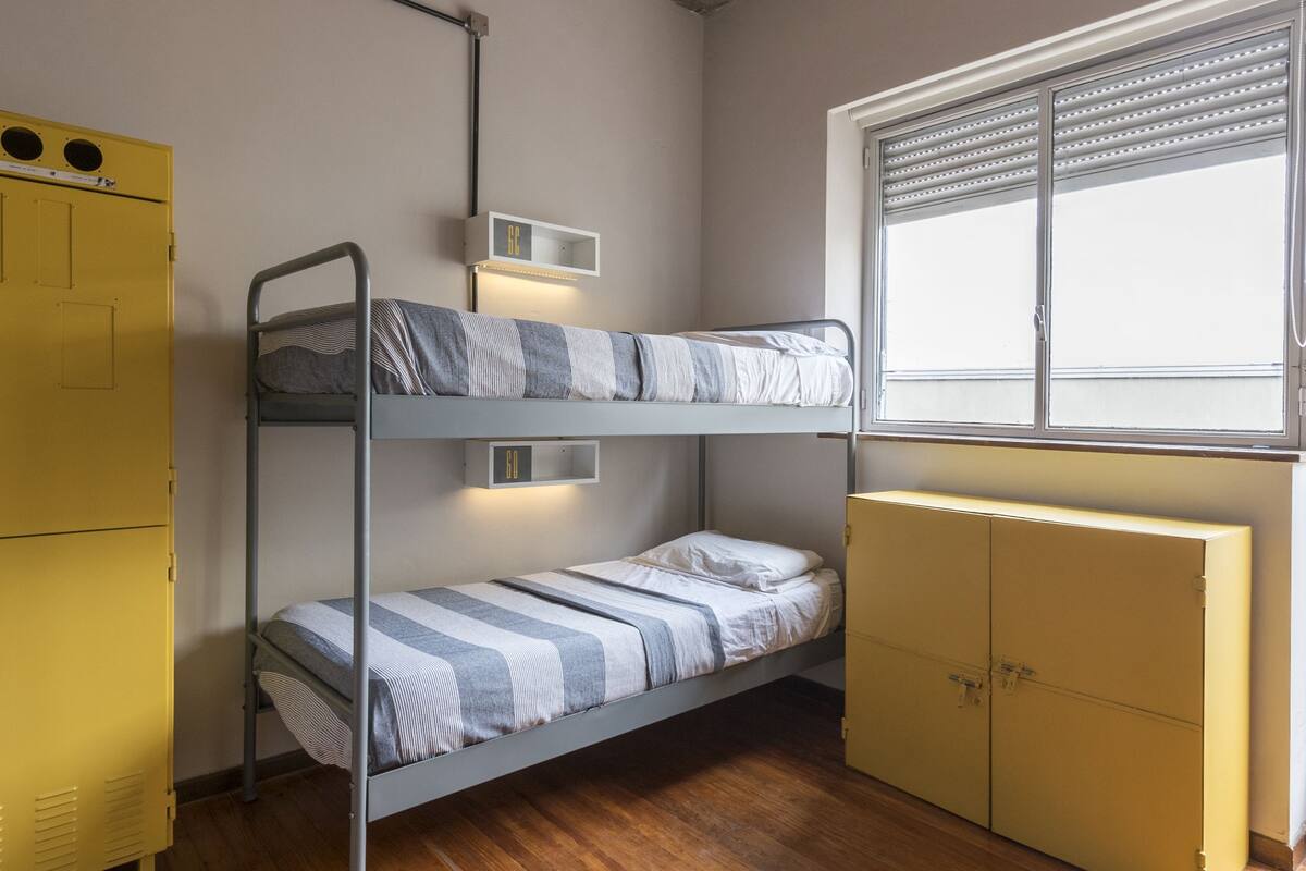 A dormitory room with bunk beds, a yellow wardrobe, and a window with blinds.