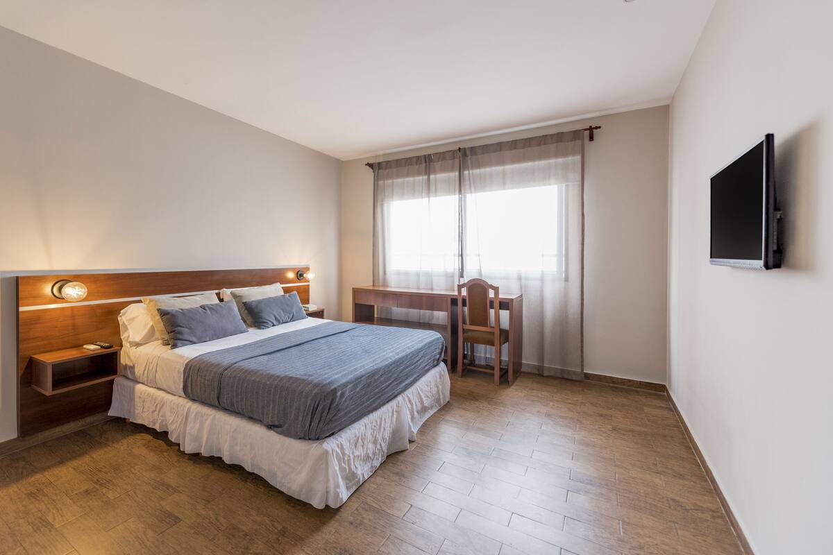 A bedroom with a bed, bedside tables, a desk, a chair, a TV, and a window with curtains.